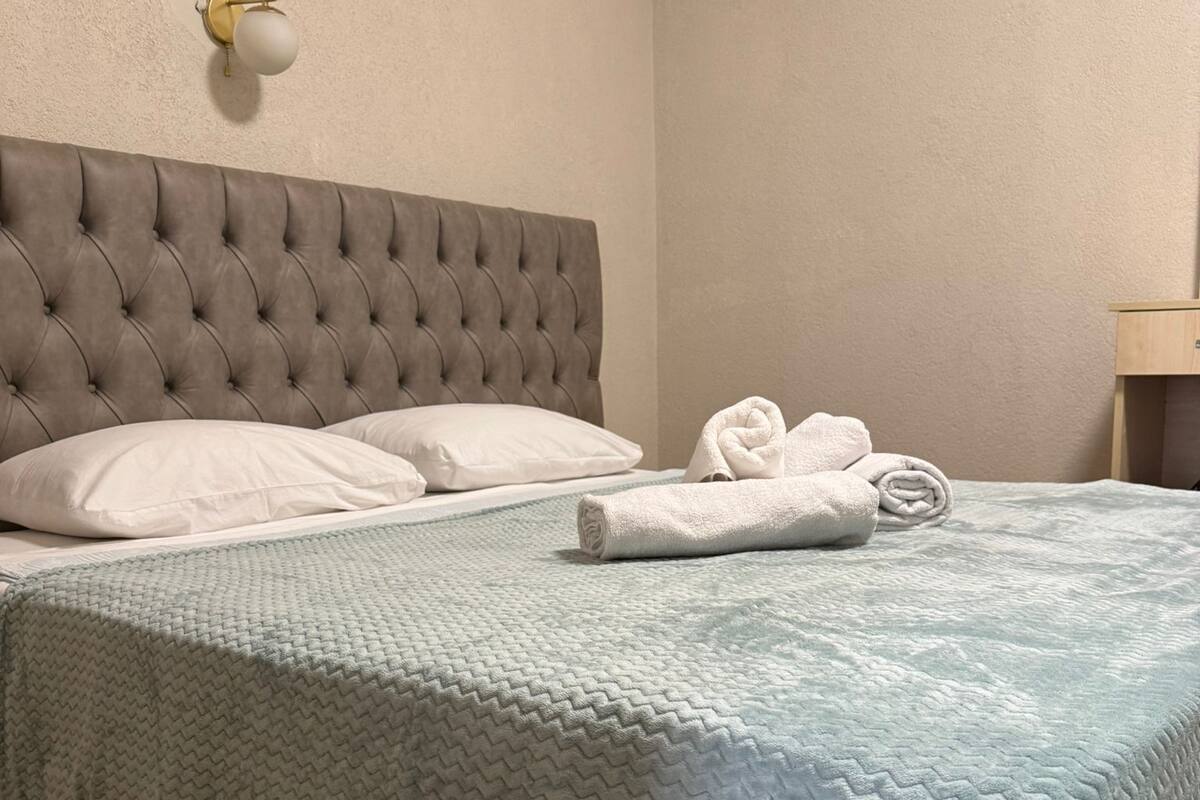 A neatly made bed with a grey headboard, a light blue quilt, and a rolled white towel on the bed.