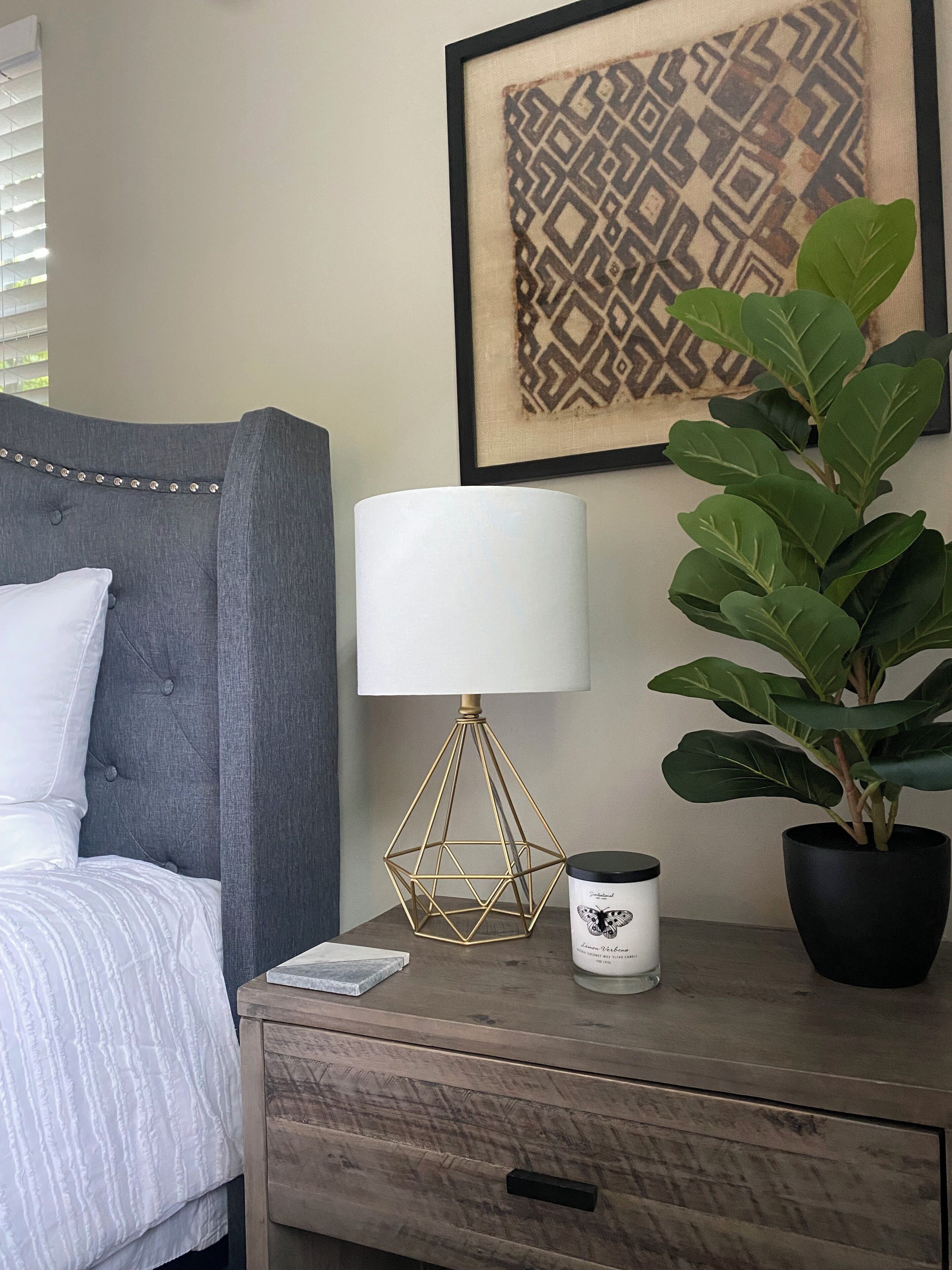 Une chambre à coucher avec un lit, une lampe de chevet, une plante en pot et une œuvre d’art encadrée accrochée au mur.
