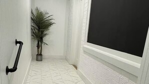 Un pasillo estrecho con paredes blancas, un muro de ladrillo blanco texturizado, una planta en maceta y una puerta negra.