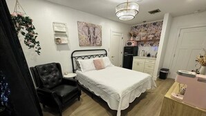 Un dormitorio con una cama, un sillón de cuero negro, una mesita de noche, una pared blanca con un estante pequeño, un refrigerador y una zona de cocina con un microondas.