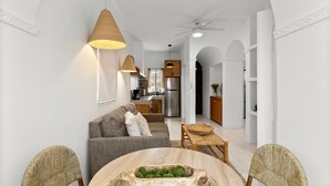 Una sala moderna con un sofá gris, una mesa de comedor de madera y una cesta tejida, y una cocina al fondo.