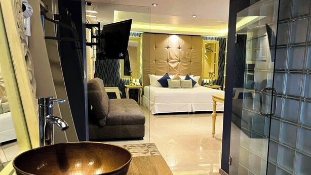 Una habitación de hotel moderna con un amplio tocador de madera, un lavamanos grande en cuenco, una cama con ropa blanca, un sillón gris y una pared divisoria de vidrio.