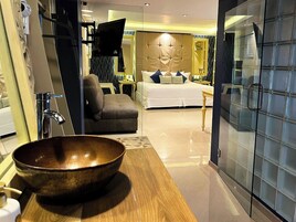 Una habitación de hotel moderna con un amplio tocador de madera, un lavamanos grande en cuenco, una cama con ropa blanca, un sillón gris y una pared divisoria de vidrio.