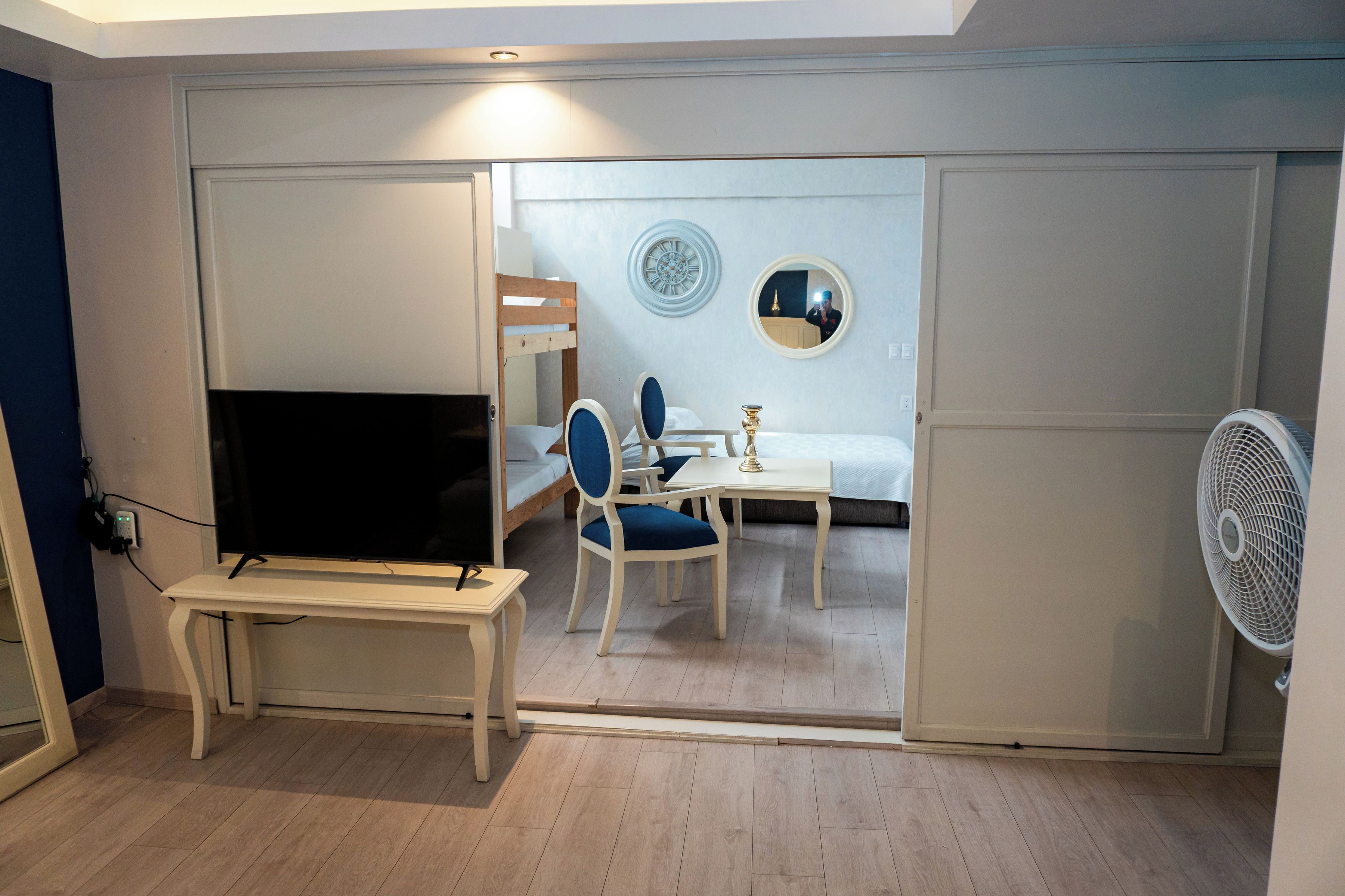 A room with a television, a dining table with chairs, a bed, and a fan.