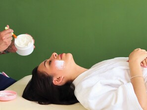 A person lying on a bed with a white bathrobe, receiving a facial treatment from another person. - Hotel Santosi (San Luis Potosí)