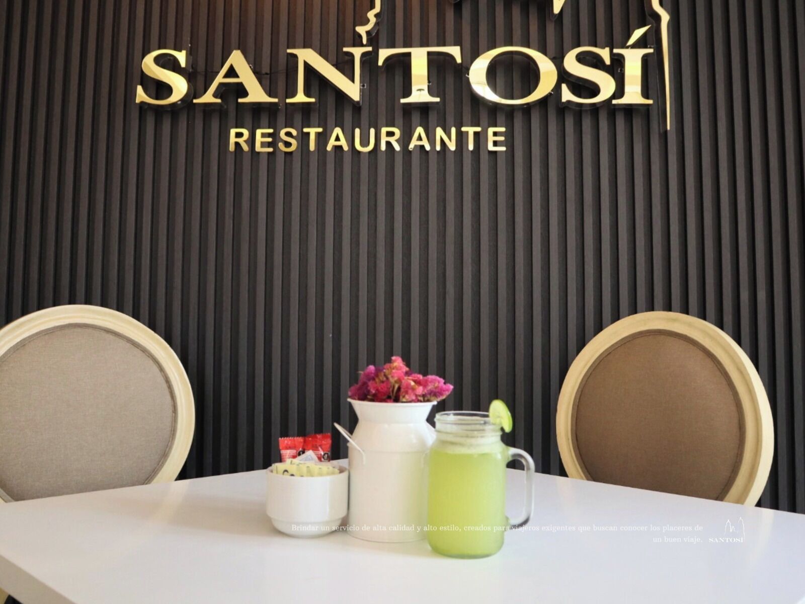 L’intérieur d’un restaurant présente un mur noir à motifs en relief et le nom « SANTOSI RESTAURANTE » en lettres dorées. On y voit deux chaises, une table blanche et un petit pichet blanc contenant une boisson verte.