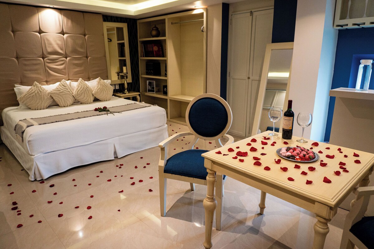 A bedroom with a large bed, a chair, a table with rose petals, and a wine bottle.