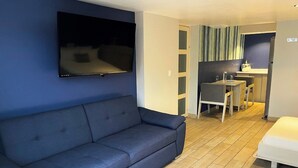 A modern living room with a blue sofa, a flat-screen TV mounted on the wall, a small dining area with a table and chairs, and a white bed. - Hotel Santosi (San Luis Potosí)