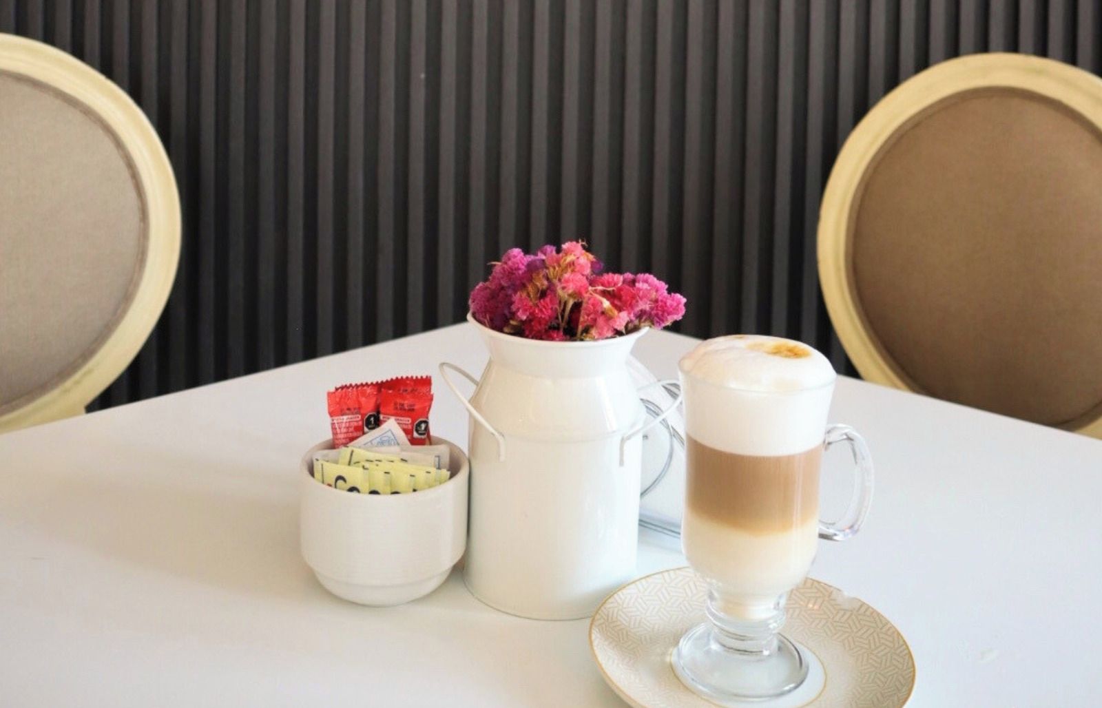 Une table avec une tasse de café, un petit vase blanc contenant des fleurs et un récipient avec de l’argent et des cartes.