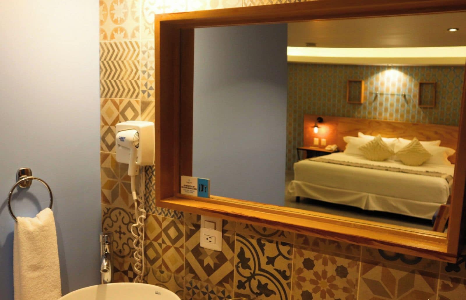 Een badkamer met een wastafel, handdoekhouder, haardroger en een spiegel die een bed weerspiegelt.