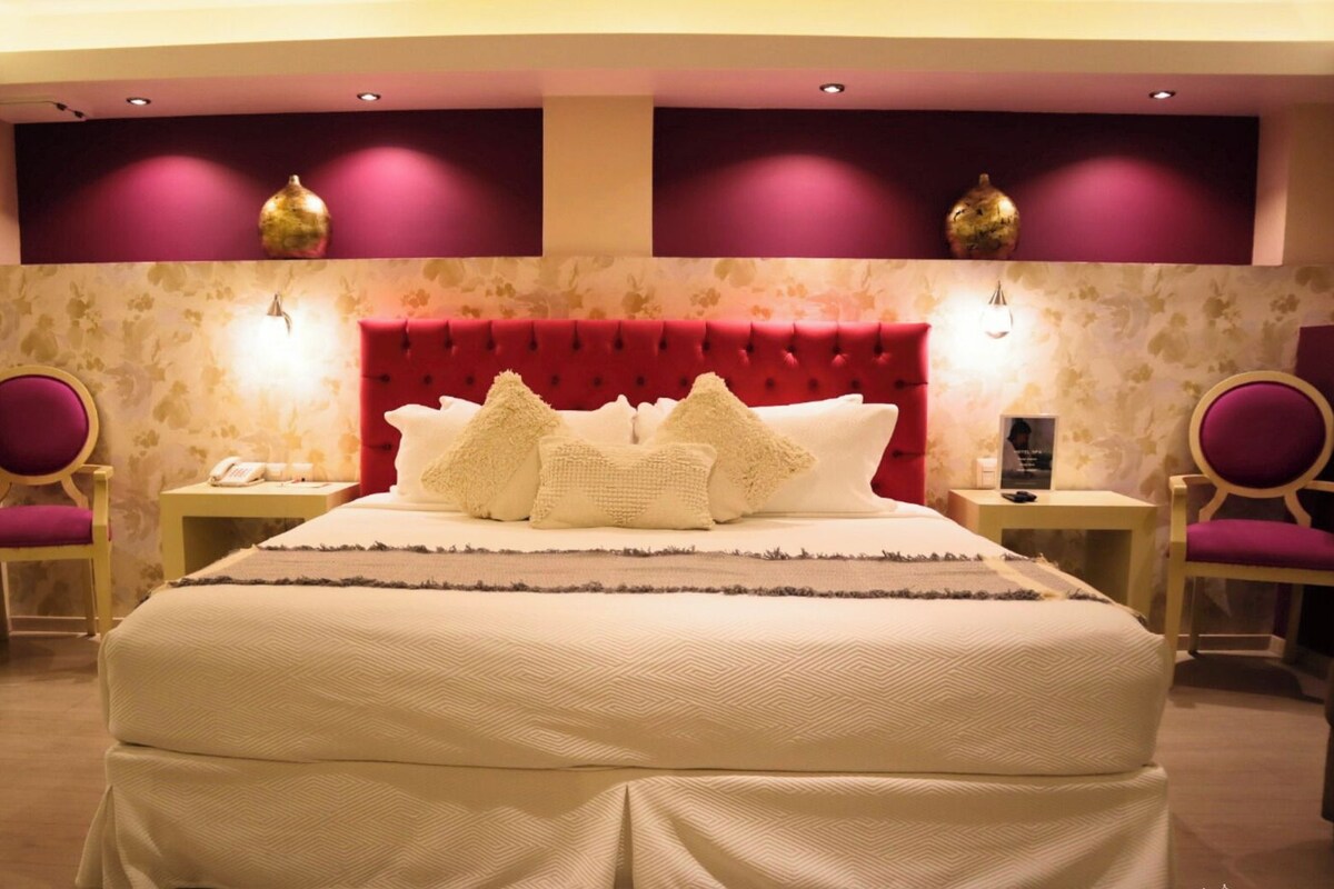 Una cama grande con cabecera roja, dos mesitas de noche y dos sillones.