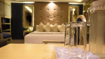 A hotel room with a neatly made bed, a bedside table with glasses and a water bottle, and a large window with curtains.