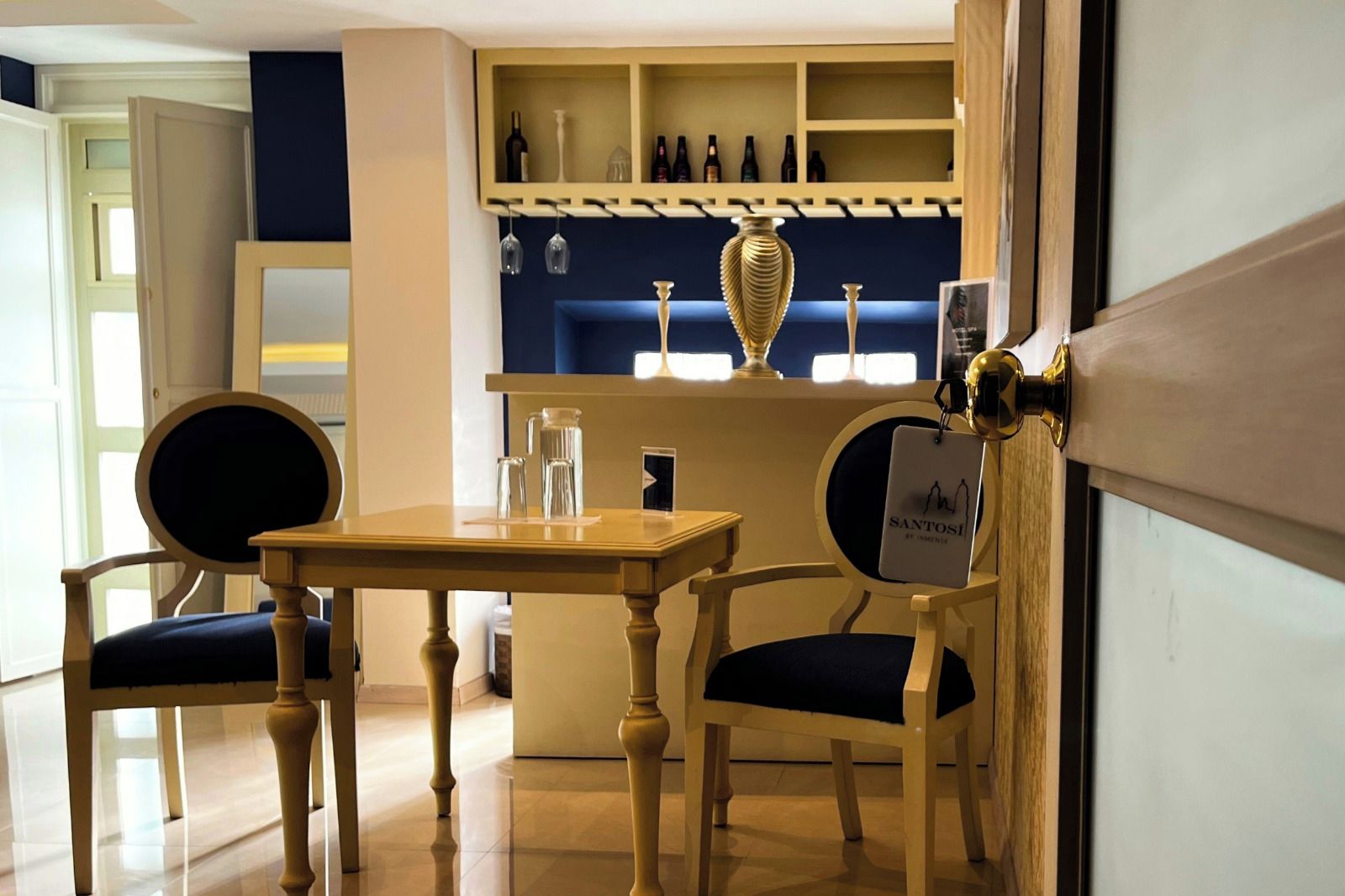 Un comedor con una mesa y sillas, una barra con estantes llenos de botellas, y un letrero que dice 'SANTOSI'.