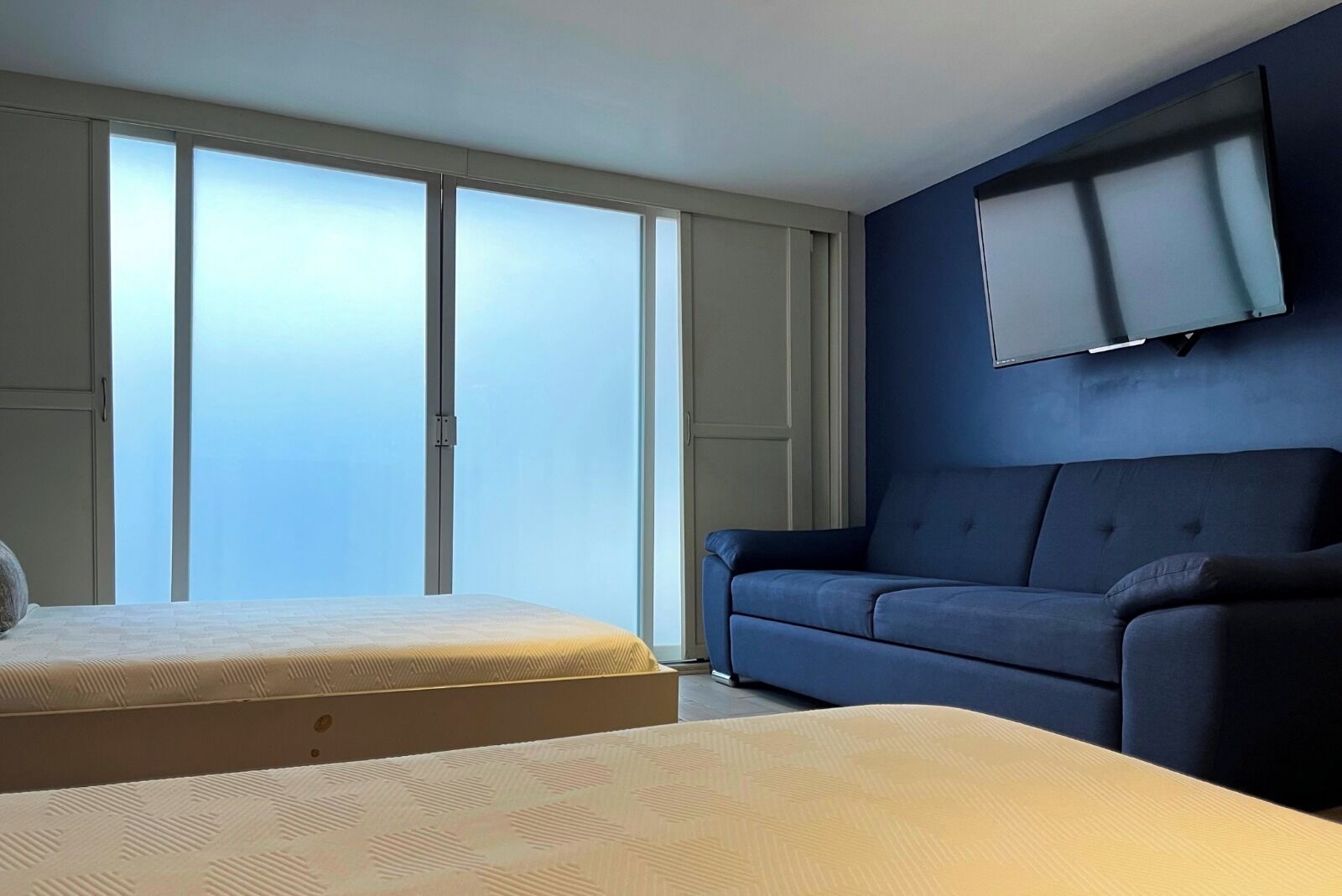 A room with a bed, a sofa, a TV mounted on the wall, and a large window.