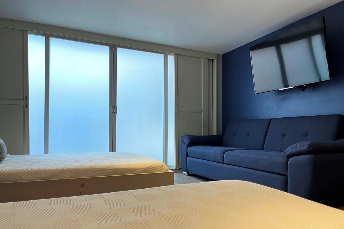 Una habitación con cama, sofá, televisor en la pared y un ventanal grande.