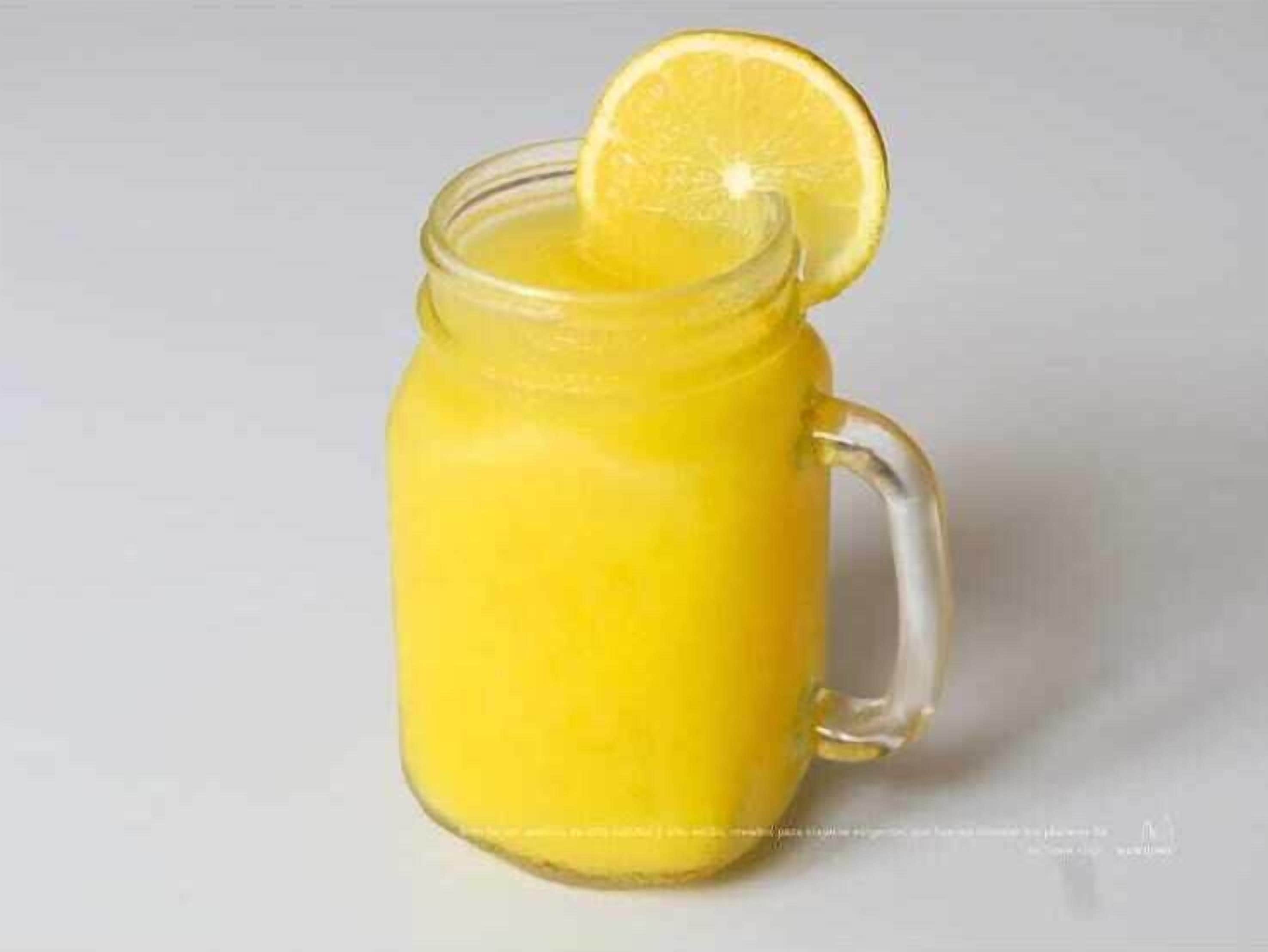 A clear glass pitcher with a handle, containing a yellow liquid, possibly a beverage, with a slice of lemon on the rim.