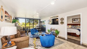 Ein Wohnzimmer mit Sofa, Sessel und Couchtisch, ein Balkon mit Blick und ein Schlafzimmer mit Bett und Fernseher.