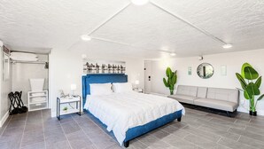 Ein modernes Hotelzimmer mit einem großen Bett, einer Couch, einem kleinen Tisch und einem Sessel. An den Wänden befinden sich Pflanzen und Kunstwerke.