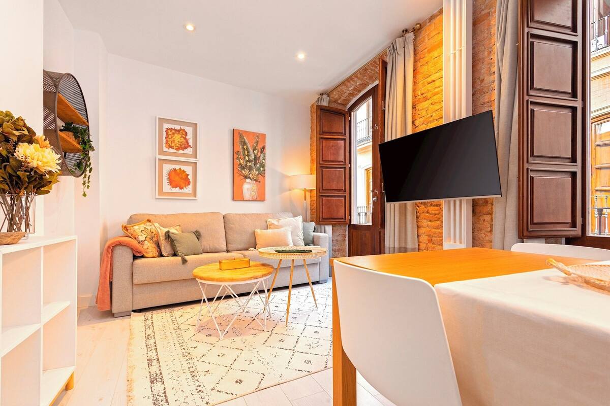 Uma sala de estar moderna com sofá, mesa de centro e televisão. Há quadros nas paredes e uma parede de tijolos ao fundo.