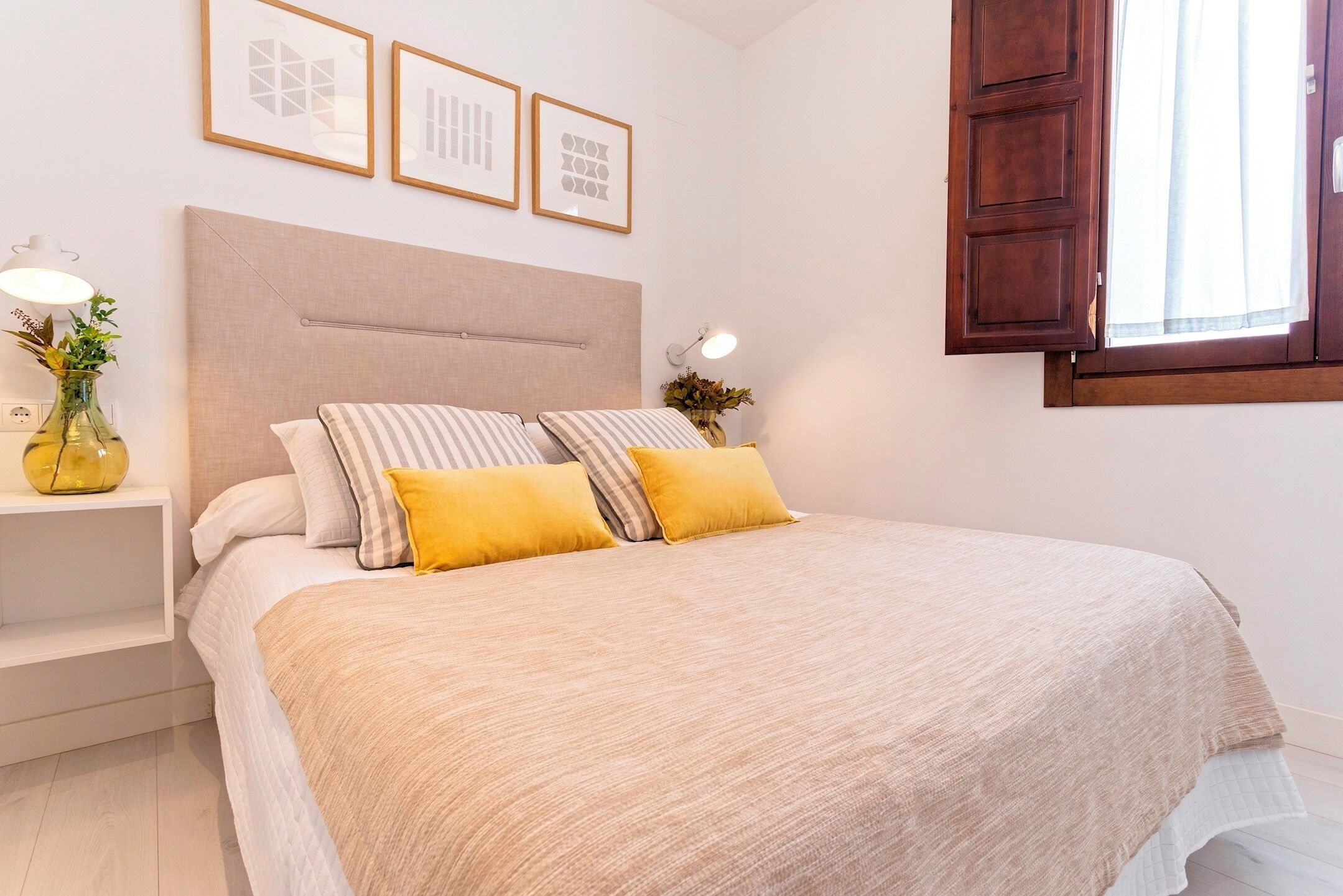 Un letto rifatto con cura, con una testiera beige, due cuscini e una coperta bianca. Sopra il letto, si vedono quadri incorniciati, e a destra una finestra con persiane in legno.