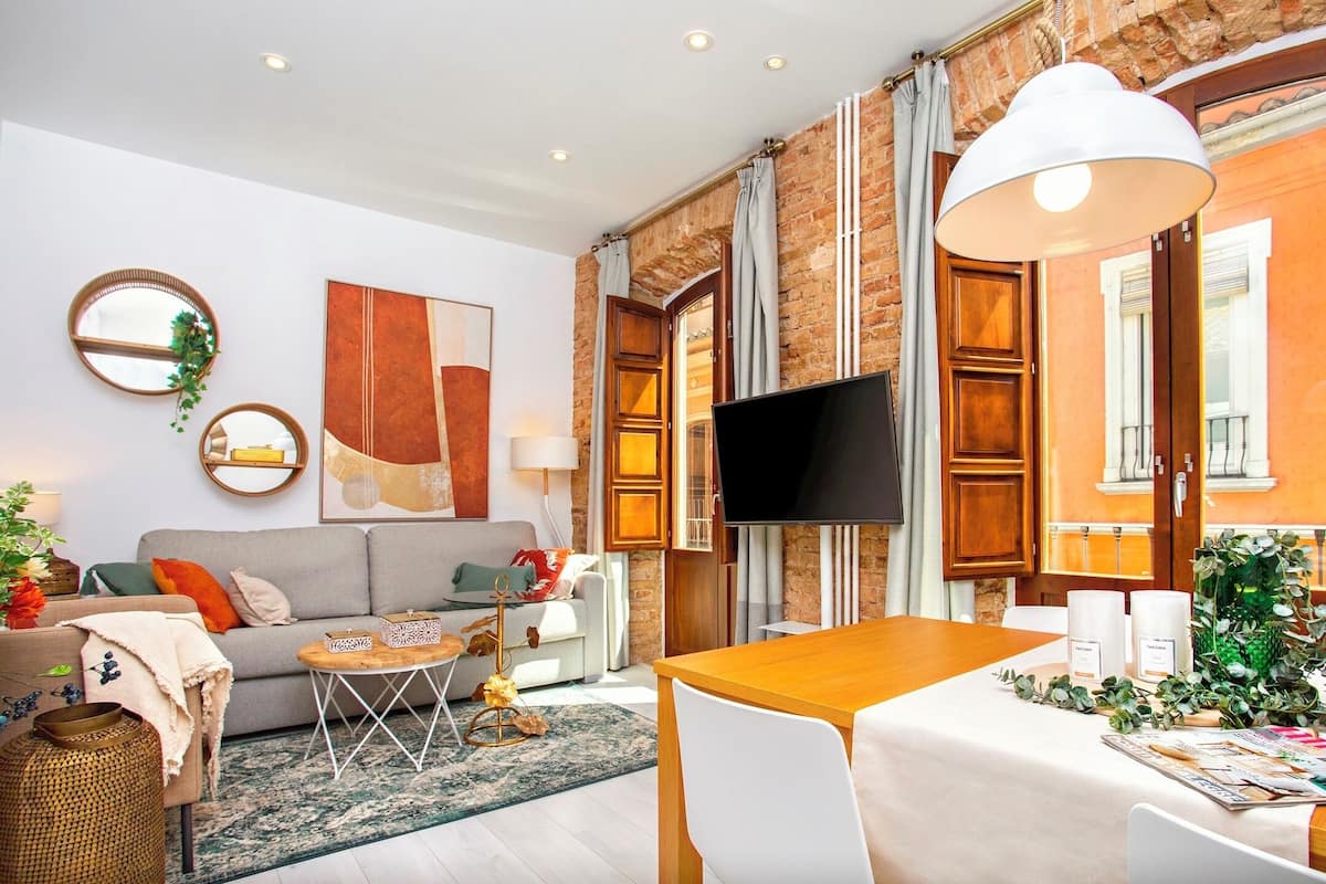 Uma sala de estar moderna com sofá, mesa de centro e televisão. Há uma área de jantar com mesa e cadeiras. O ambiente possui uma parede de tijolos e amplas janelas.