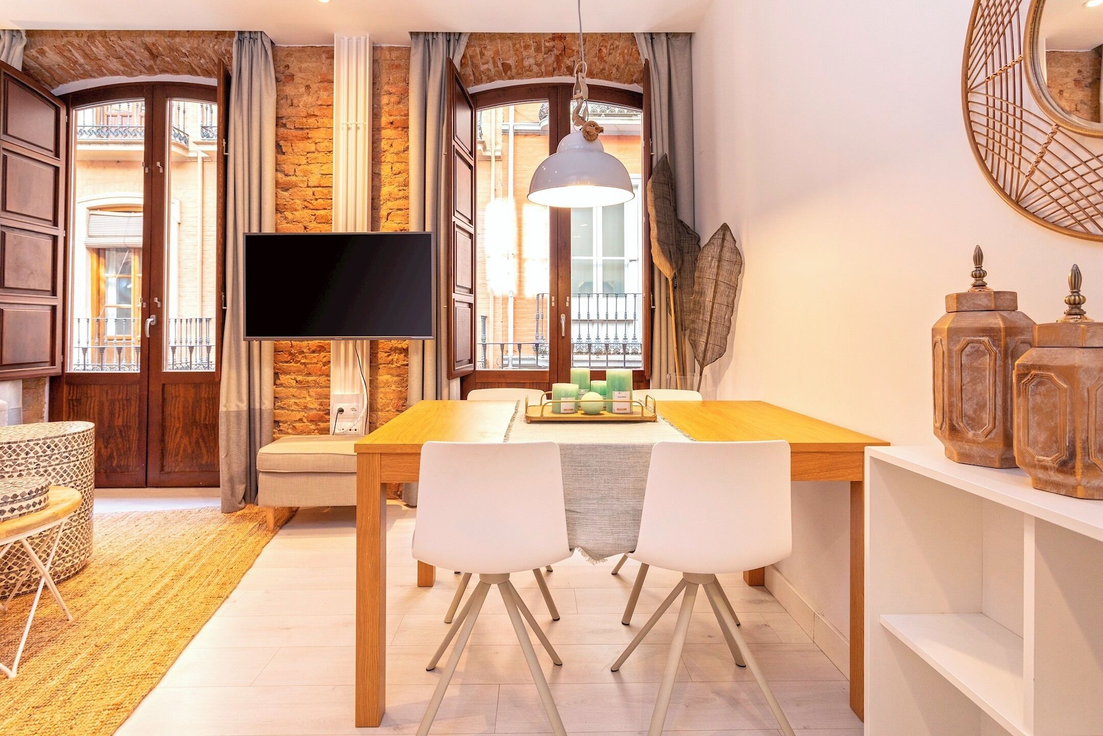 A dining area with a wooden table, two white chairs, a TV mounted on the wall, and a view of a balcony through large windows.