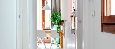 Un pasillo estrecho con piso estampado que lleva a un comedor con sillas y una mesa, una planta en maceta y un refrigerador.