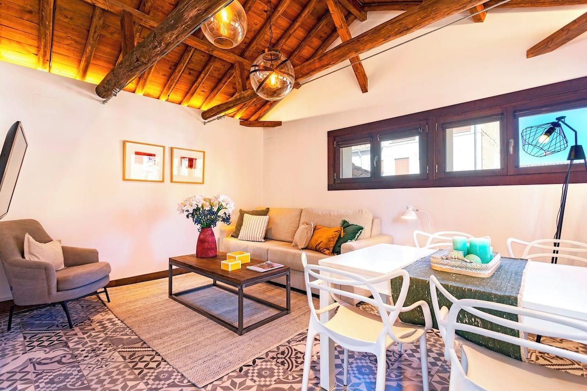 Uma sala de estar acolhedora com teto de madeira, um sofá, uma mesa de centro e um espaço de jantar com cadeiras brancas.