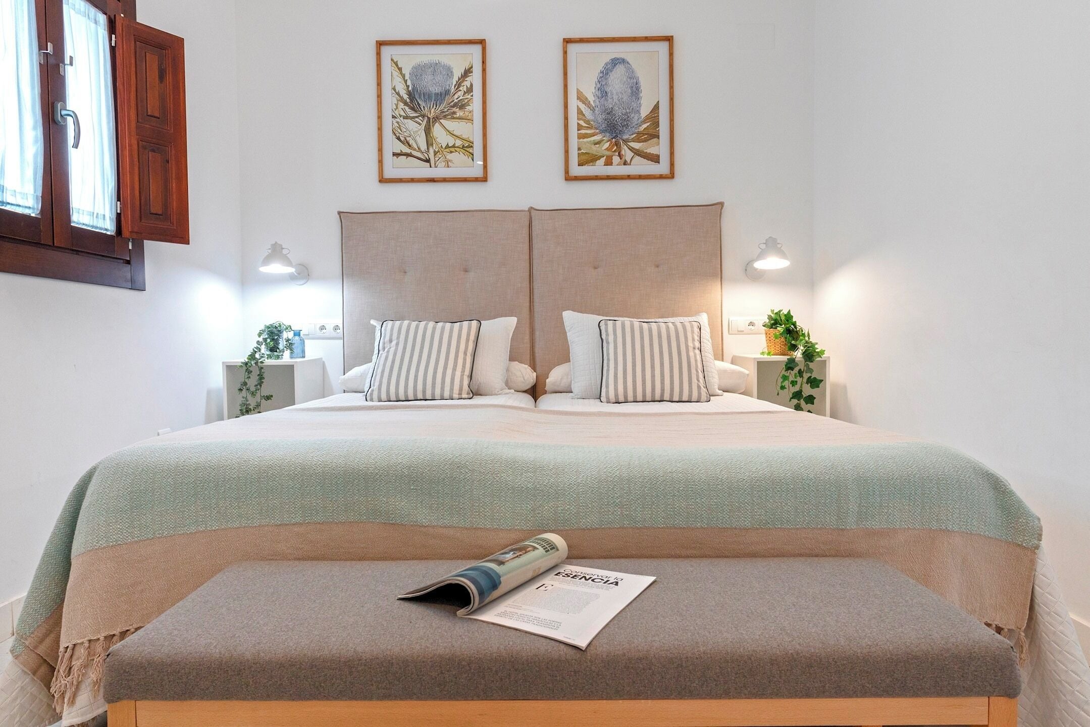 A neatly made bed with a light green bedspread, two pillows, and a magazine on a bench. There are two wall-mounted lamps, a window with wooden shutters, and two framed botanical prints above the bed.