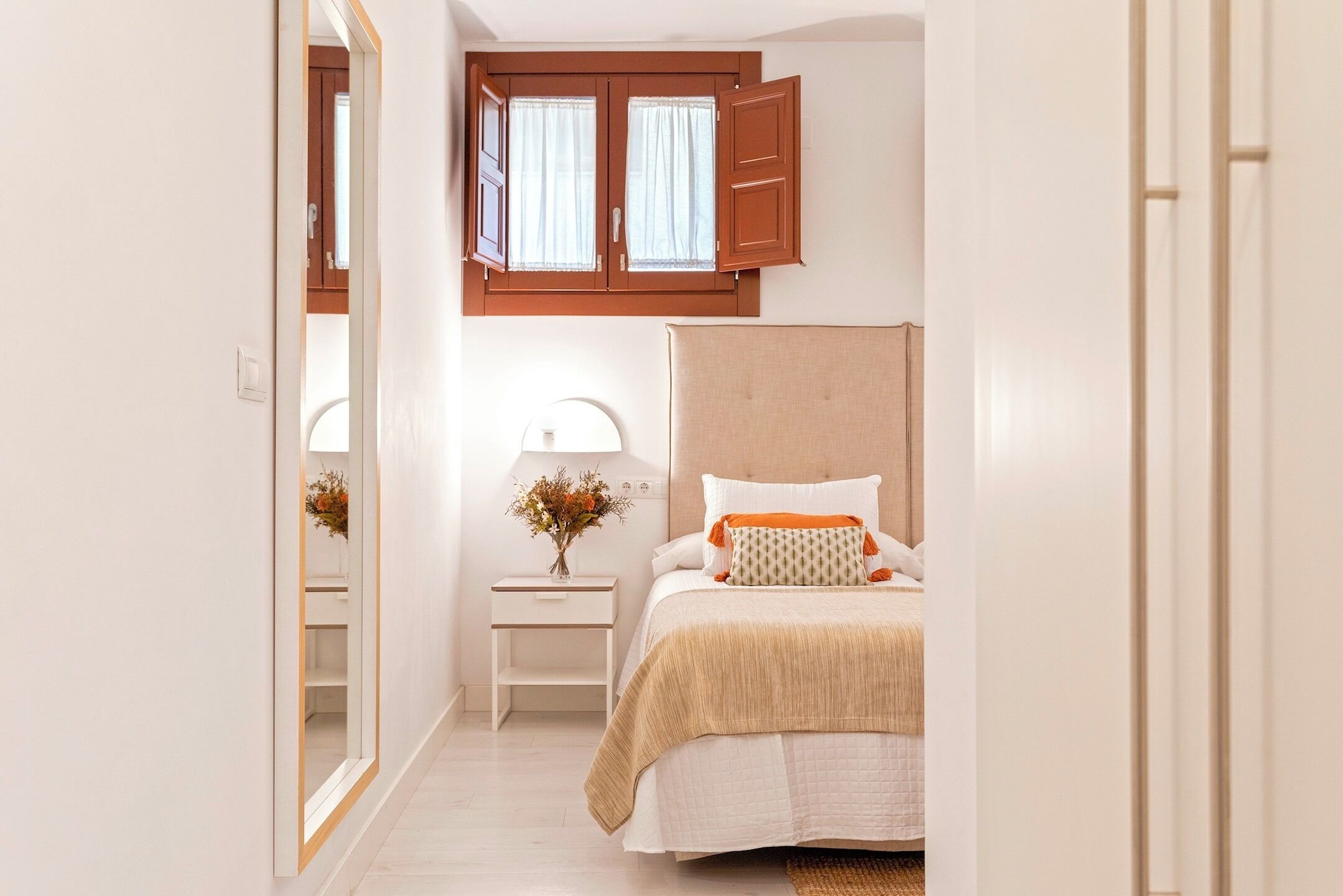 A bedroom with a bed, nightstand, mirror, window, and wooden wardrobe.