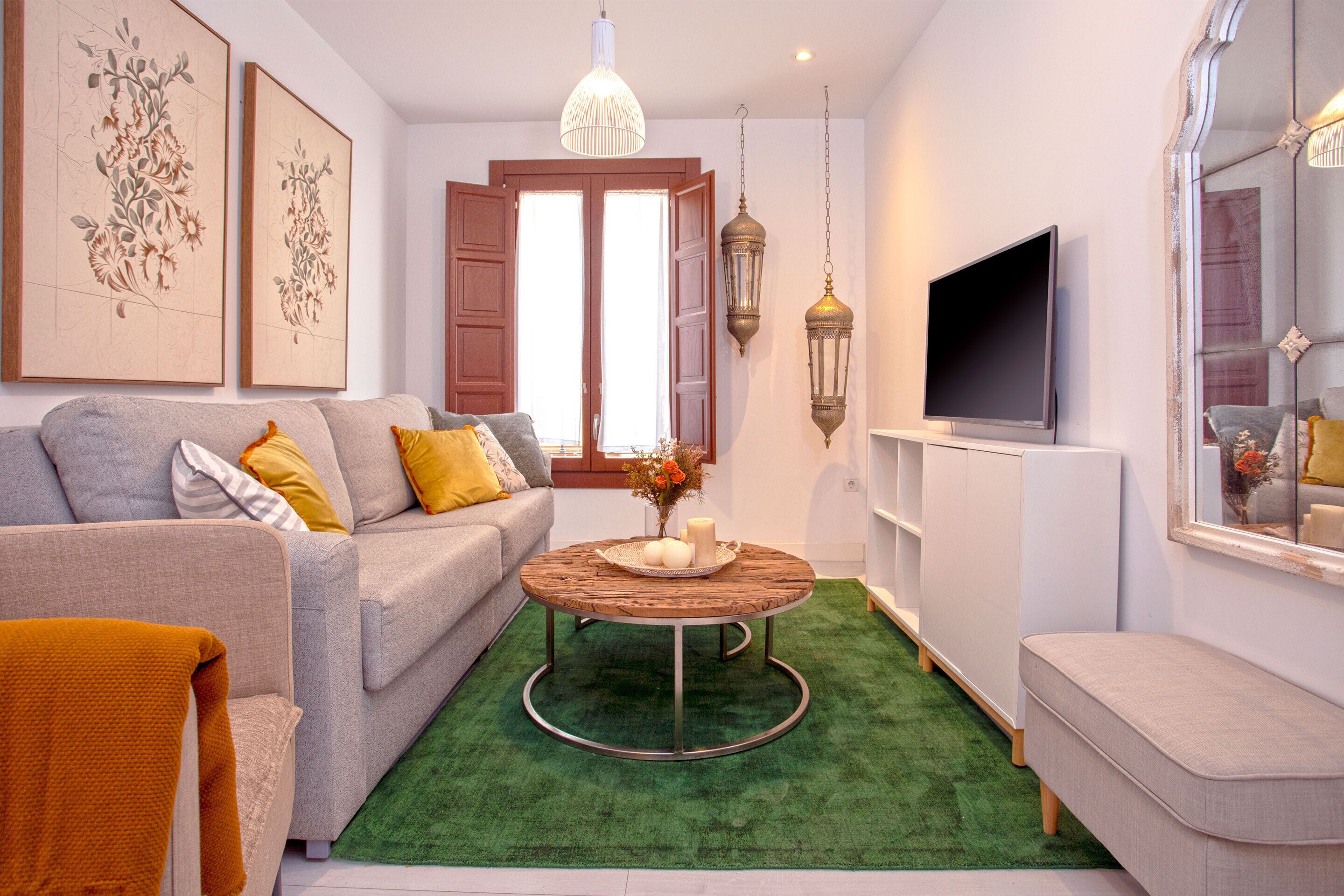 A modern living room with a grey sofa, a round coffee table with a vase, a flat-screen TV, and a green rug.
