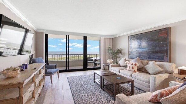 A spacious living room with a large flat-screen TV, a sofa set, a coffee table, and a patterned rug. A balcony with a view of the ocean is visible through the glass doors.