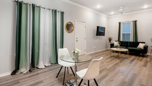 A modern living room with a glass table, white chairs, a green sofa, and a ceiling fan. - 1BR The Raven Flats B! Hermann Park! Med Center! (Houston)