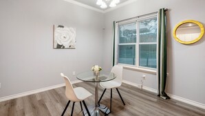 A dining area with a glass table, two white chairs, a vase with flowers, a painting on the wall, a window with blinds, and a round mirror. - 1BR The Raven Flats B! Hermann Park! Med Center! (Houston)
