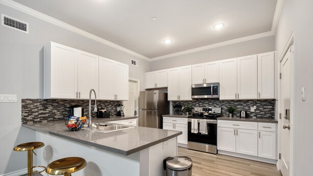 A modern kitchen with white cabinets, a granite countertop, stainless steel appliances, and gold bar stools. - 1BR The Raven Flats B! Hermann Park! Med Center! (Houston)
