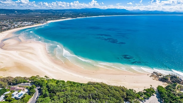 Luftaufnahme einer Küstenlandschaft mit Sandstrand, klarem blauen Wasser und dichter Vegetation.