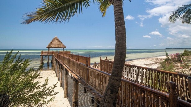 A wooden deck leading to a thatched-roof structure overlooking a beach with clear blue water. - Zanzibar Beachfront Villa 1012 (Zanzibar)