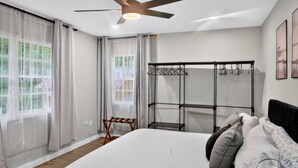 Un dormitorio con cama, ventilador de techo, ventana con cortinas y un estante metálico en la pared.