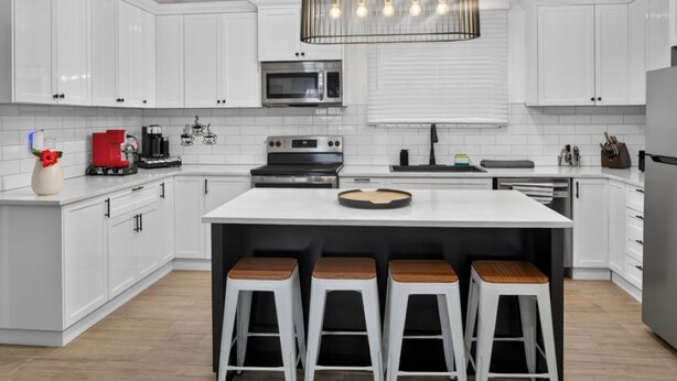 Una cocina moderna con muebles blancos, una isla central con taburetes, una lámpara colgante y un microondas sobre la isla.
