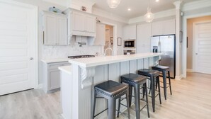 A modern kitchen with white cabinetry, a central island with stools, stainless steel appliances, and pendant lighting. - Coastal Comfort | 4BR Beach Retreat with Beach Views +  ShortWalk to Shore! (Pass Christian)