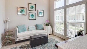 A modern living room with a white sofa, a black ottoman, and a patterned rug. There are four framed artworks on the wall and a small table with a potted plant. - Coastal Comfort | 4BR Beach Retreat with Beach Views +  ShortWalk to Shore! (Pass Christian)