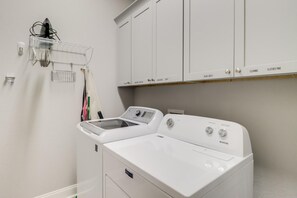 A laundry area with a front-loading washer and dryer, a drying rack, and a cabinet above with labeled compartments for cleaning and clothes. - Coastal Comfort | 4BR Beach Retreat with Beach Views +  ShortWalk to Shore! (Pass Christian)