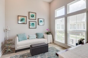 Ein modernes Wohnzimmer mit einem weißen Sofa, einem schwarzen Hocker und einem gemusterten Teppich. Vier gerahmte Kunstwerke sind an der Wand und ein kleiner Tisch mit einer Topfpflanze zu sehen.