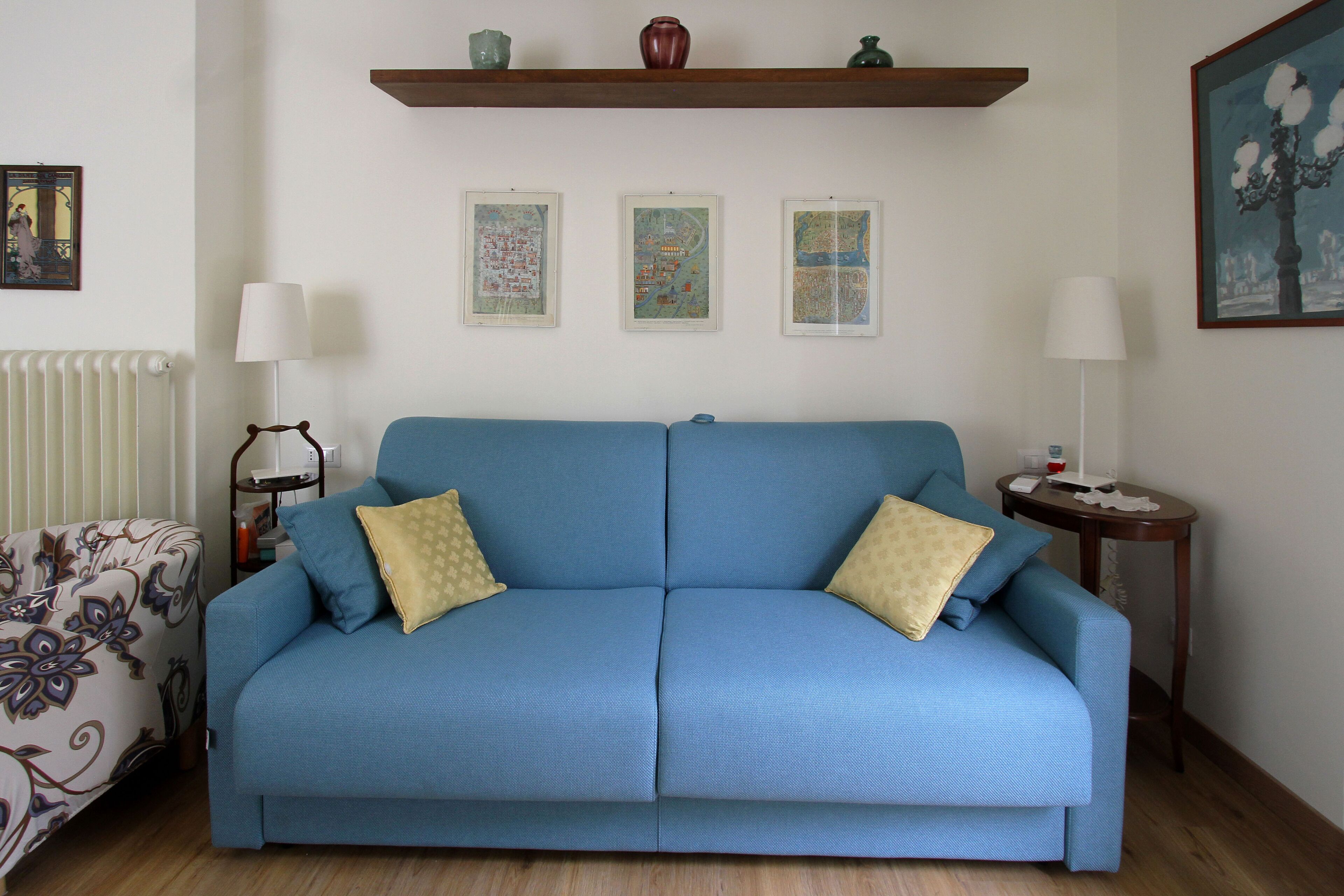 Un sofá azul con dos cojines amarillos, un sillón estampado, una mesita con una lámpara y tres mapas enmarcados en la pared.