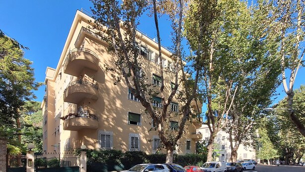Un edificio de varios pisos con balcones, coches aparcados y árboles.