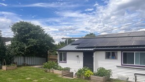 A single-story house with a solar panel roof, surrounded by a well-maintained lawn and garden beds.