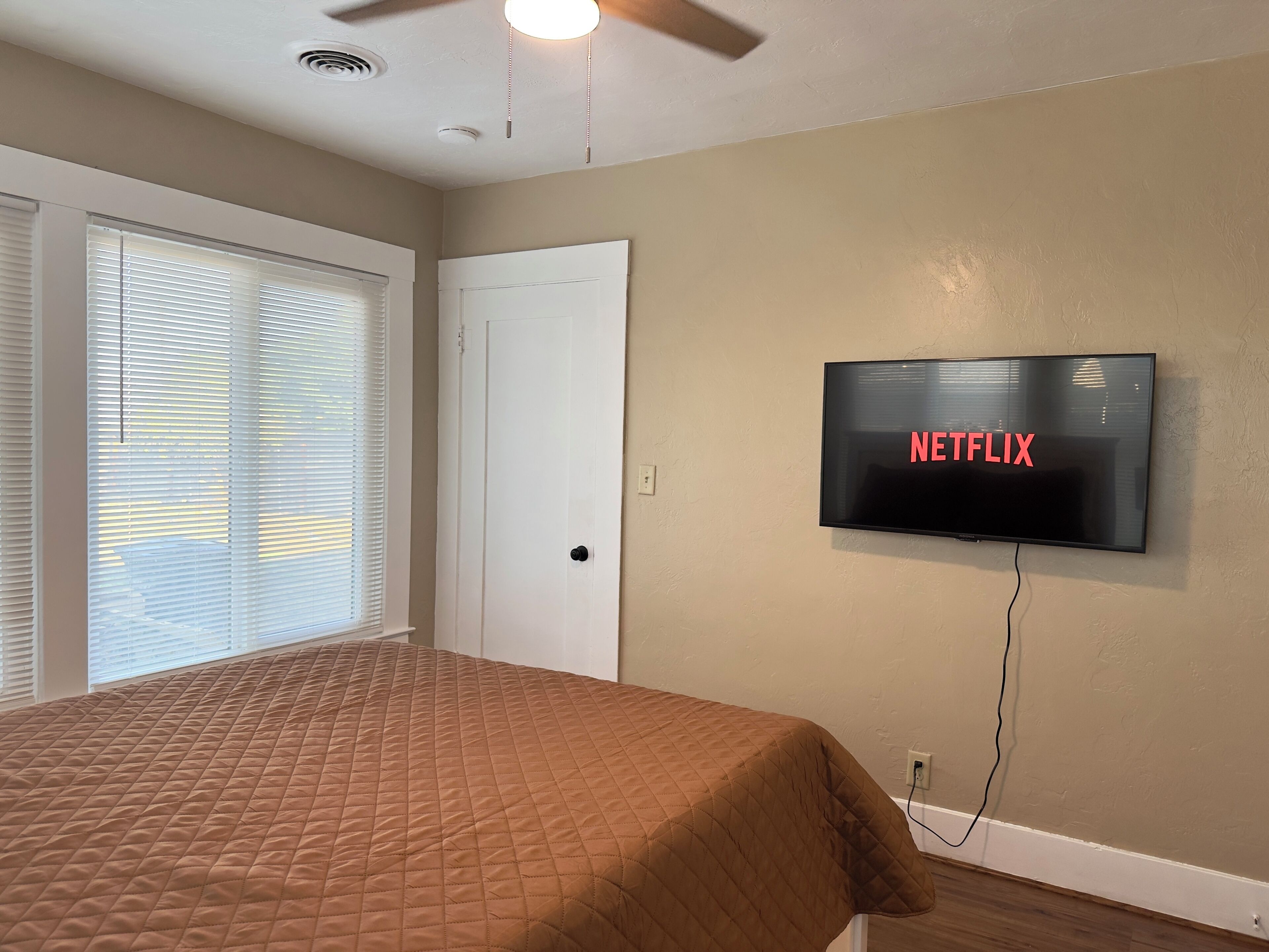 A bedroom with a bed, a door, a wall-mounted TV displaying 'NETFLIX', and a ceiling fan.