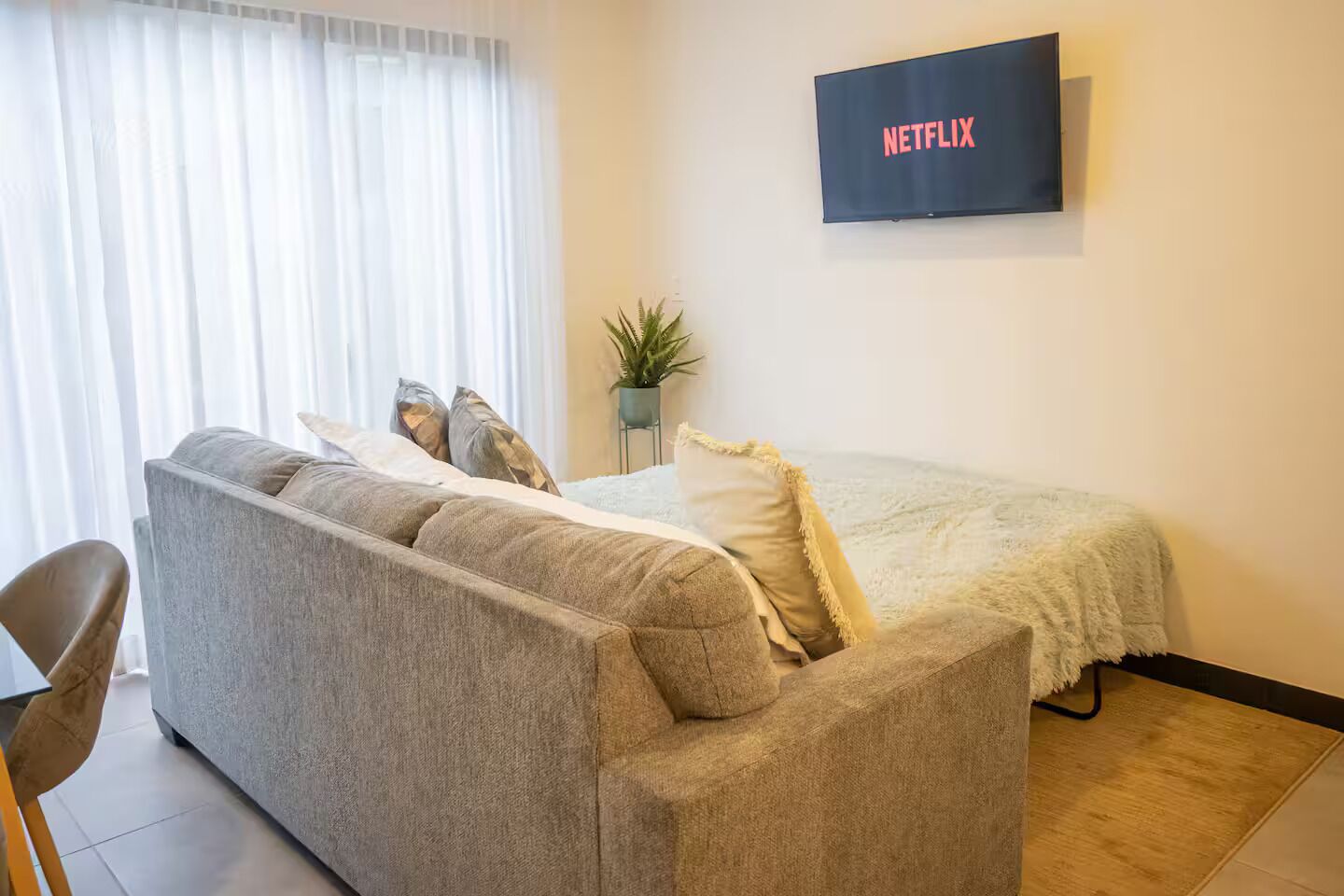 Een moderne kamer met een grijze bank, een bed met een witte dekbedovertrek, een kleine potplant en een tv aan de muur waarop het Netflix-logo te zien is.