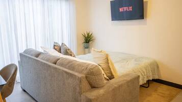 Een moderne kamer met een grijze bank, een bed met een witte dekbedovertrek, een kleine potplant en een tv aan de muur waarop het Netflix-logo te zien is.