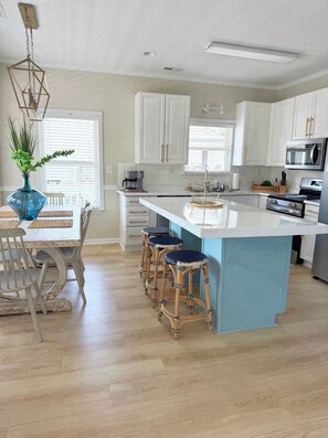 A modern kitchen with white cabinets, a central island with stools, a dining table with chairs, and a hanging light fixture.
